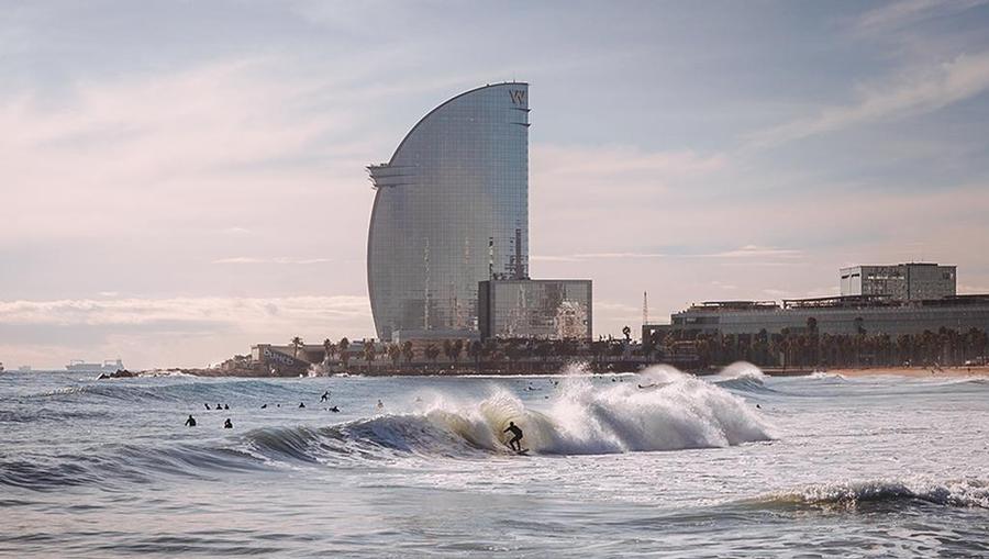 The W hotel on the coast of Barcelona with surfers riding waves on the beach below.